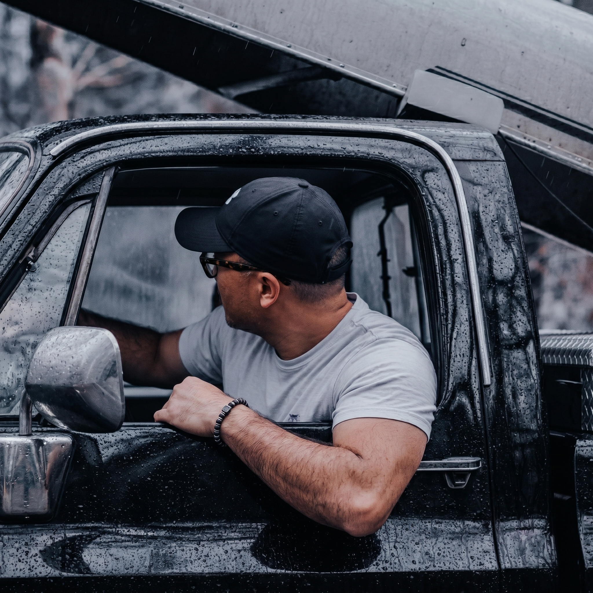 Man sitting inside a wet truck with the hood open, wearing a cap and sunglasses.