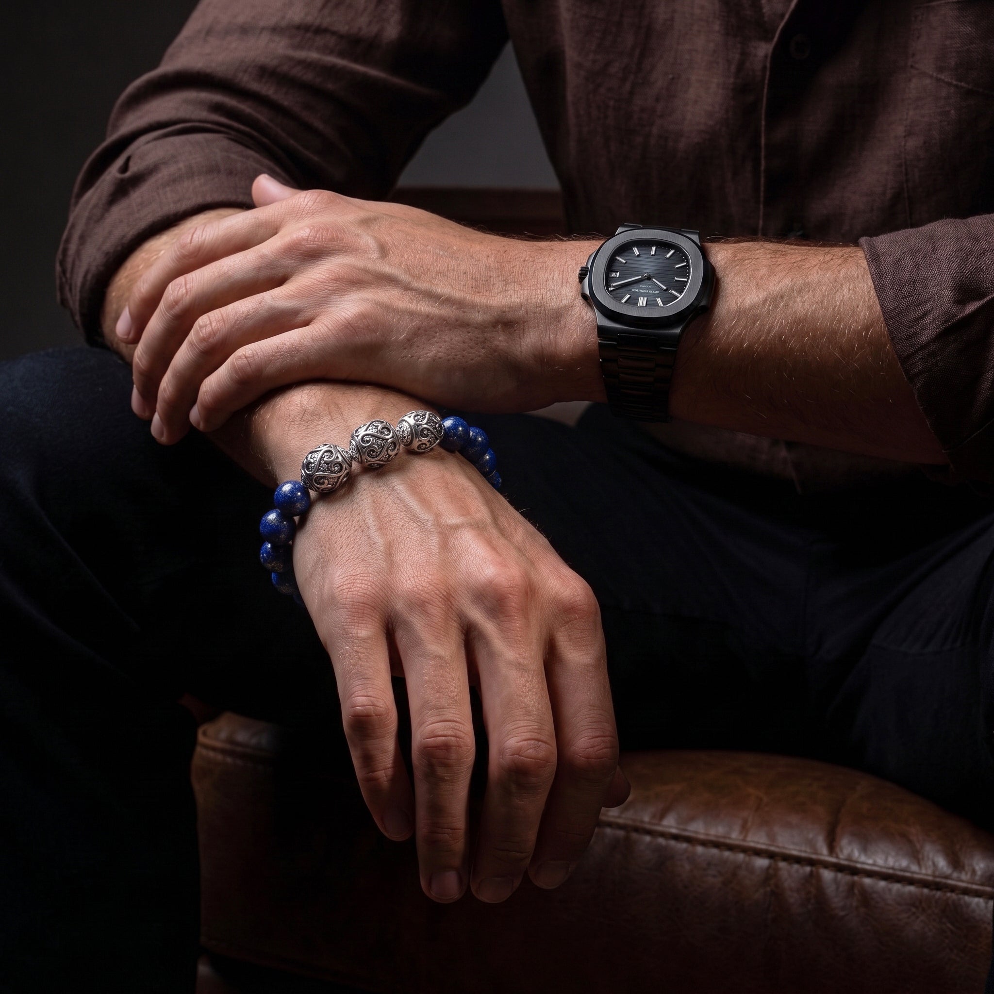 A close-up lifestyle shot of a man wearing a dark brown shirt and black pants, showcasing a dark wood watch on his wrist and a blue beaded bracelet with silver decorative elements on his other wrist, while seated in a brown leather armchair.
