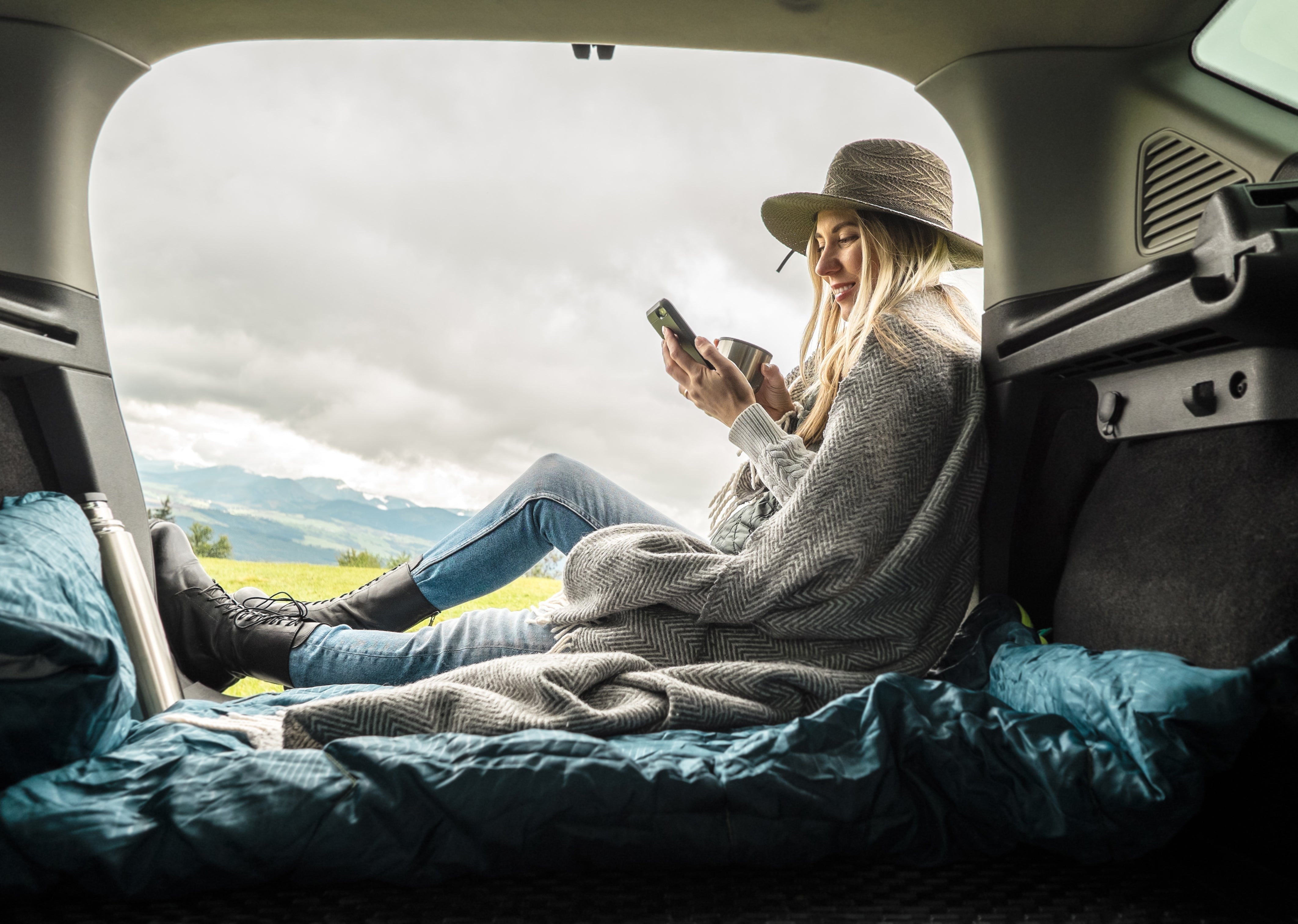 Woman sitting in the back of a car with an open trunk, looking at her phone.