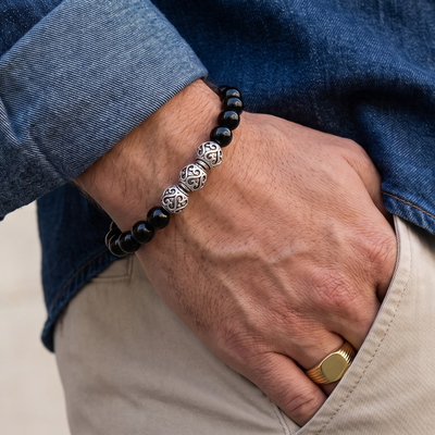 A close-up of a man's hand wearing a black beaded bracelet with three silver-toned circular charms, styled with a blue denim shirt and beige trousers.