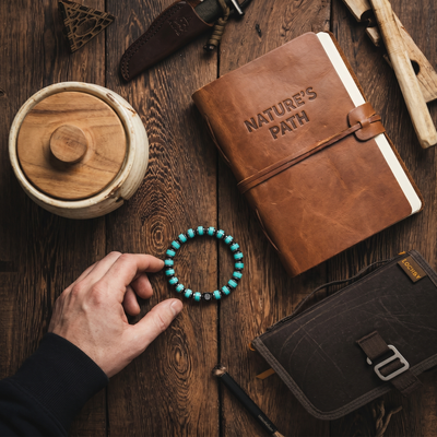 Brown leather notebook with 'Nature's Path' embossed, hand holding turquoise bracelet on wooden surface