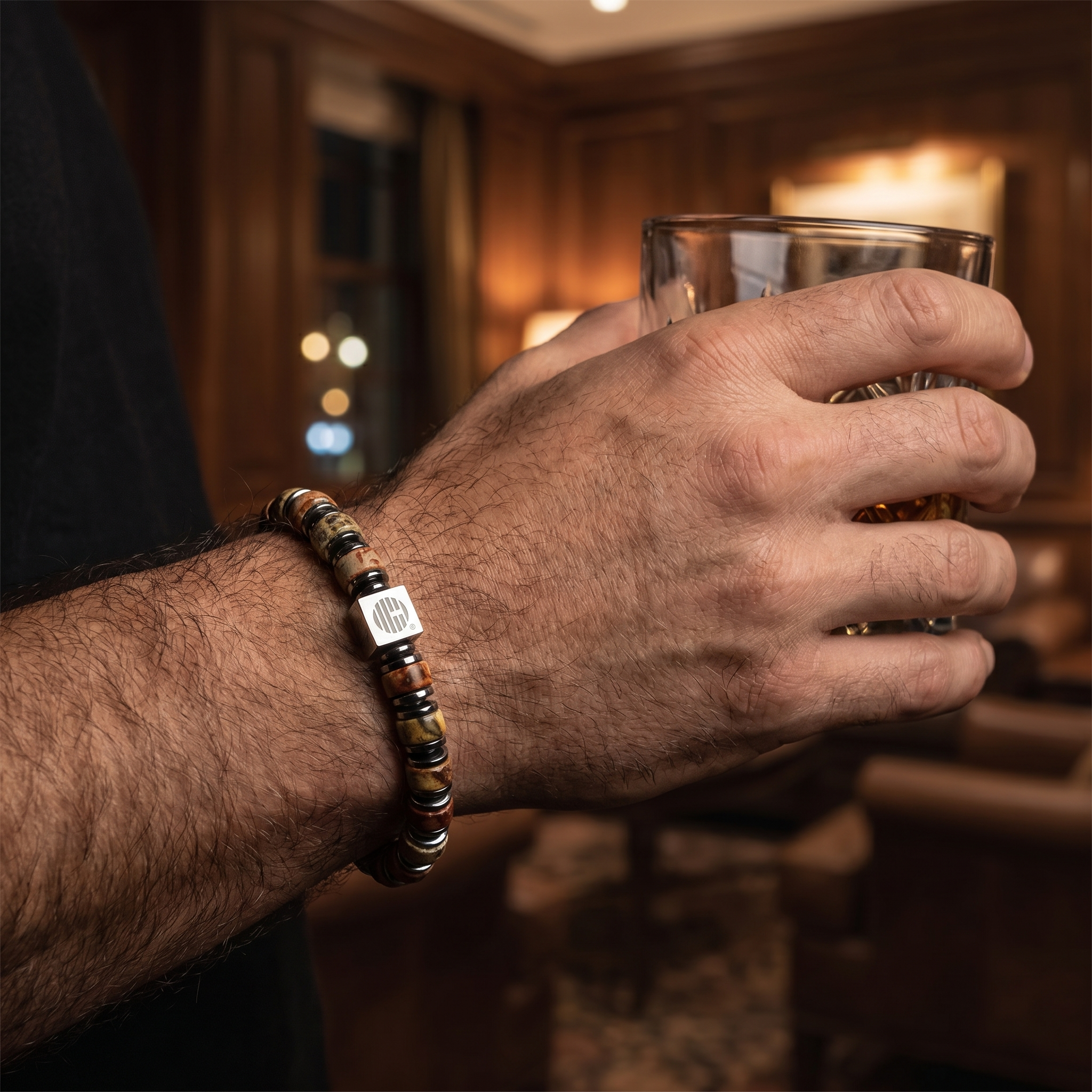 Hand holding a glass with a bracelet featuring a logo, in a warm-toned indoor setting.